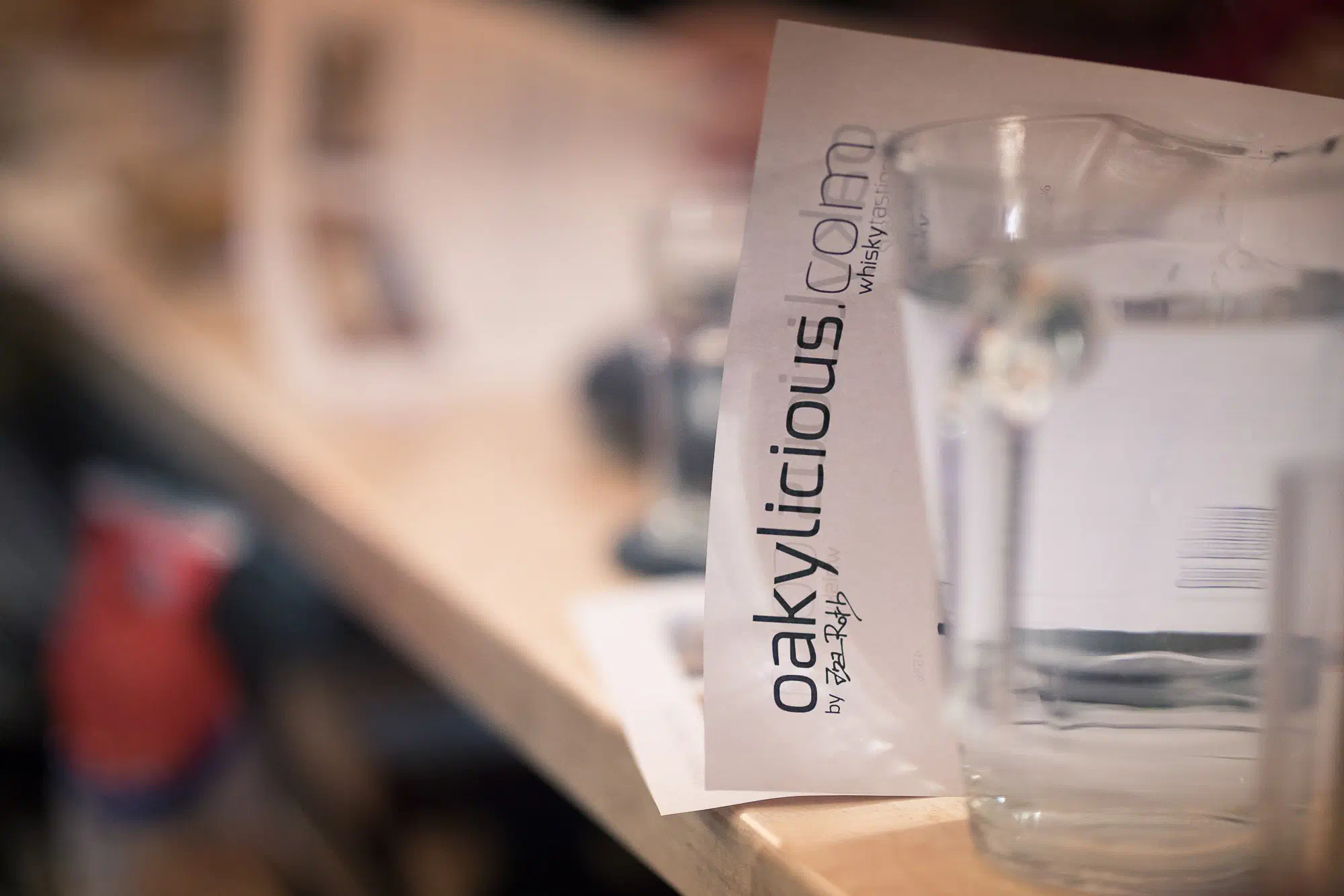 Eine Karte mit dem Schriftzug oakylicious.com lehnt auf einer Holztheke neben einer Glaskaraffe mit Wasser