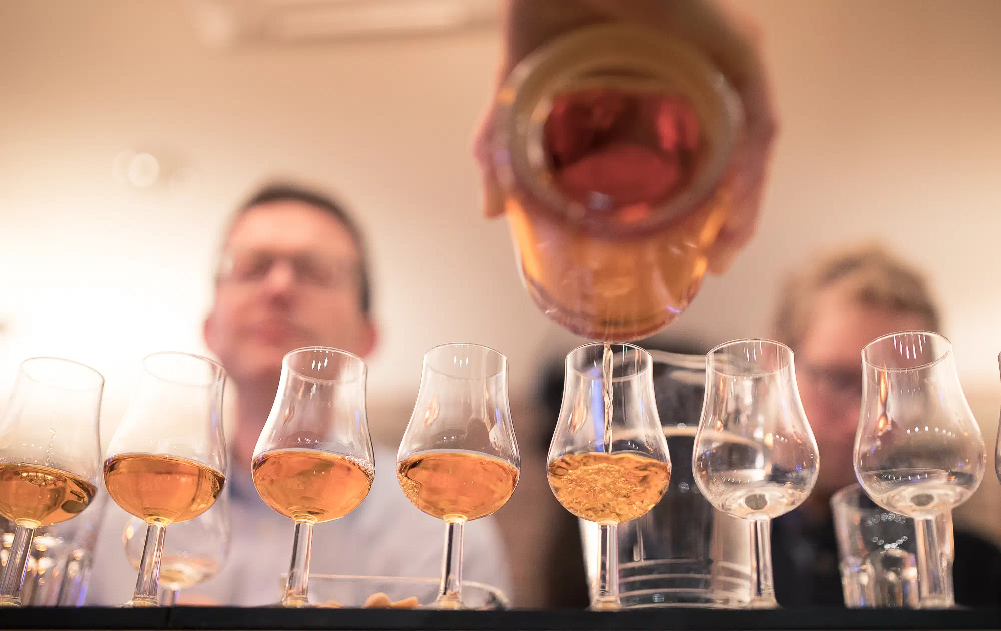 Eine Reihe Nosing-Gläser auf dem Tresen, in eins wird Whisky aus der Flasche eingeschenkt, dahinter zwei Gäste unscharf