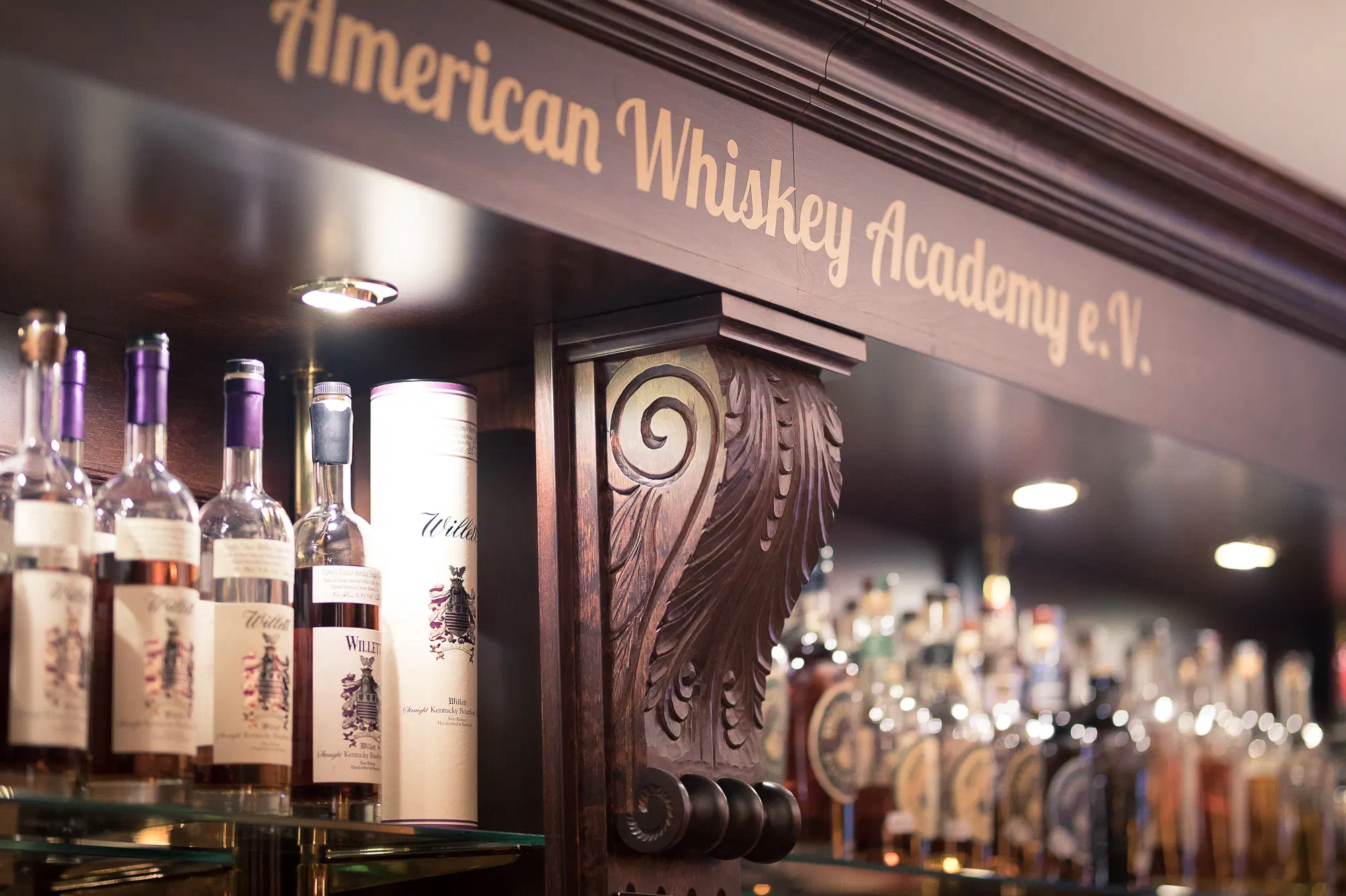 Flaschen Willett Bourbon im Regal unter einem geschnitzten Holzbalken mit dem Schriftzug American Whiskey Academy e.V.