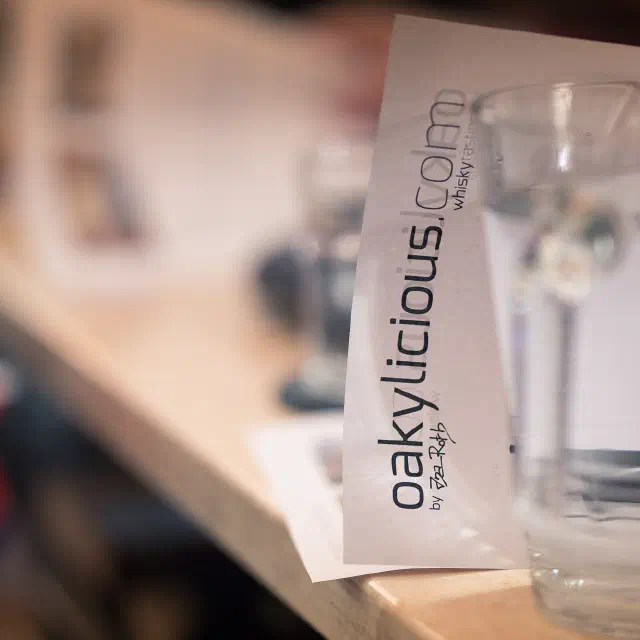 Eine Tasting-Karte mit dem Schriftzug oakylicious.com lehnt auf dem Tresen neben einer Karaffe mit Wasser