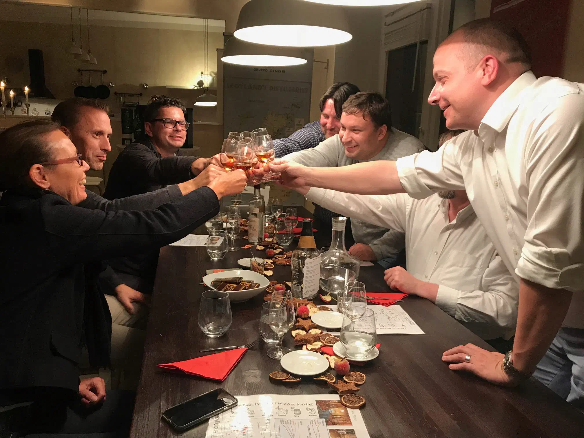 Gäste an einer gedeckten Tafel heben lachend ihre Whiskygläser zum Anstoßen, im Hintergrund eine Karte der schottischen Brennereien