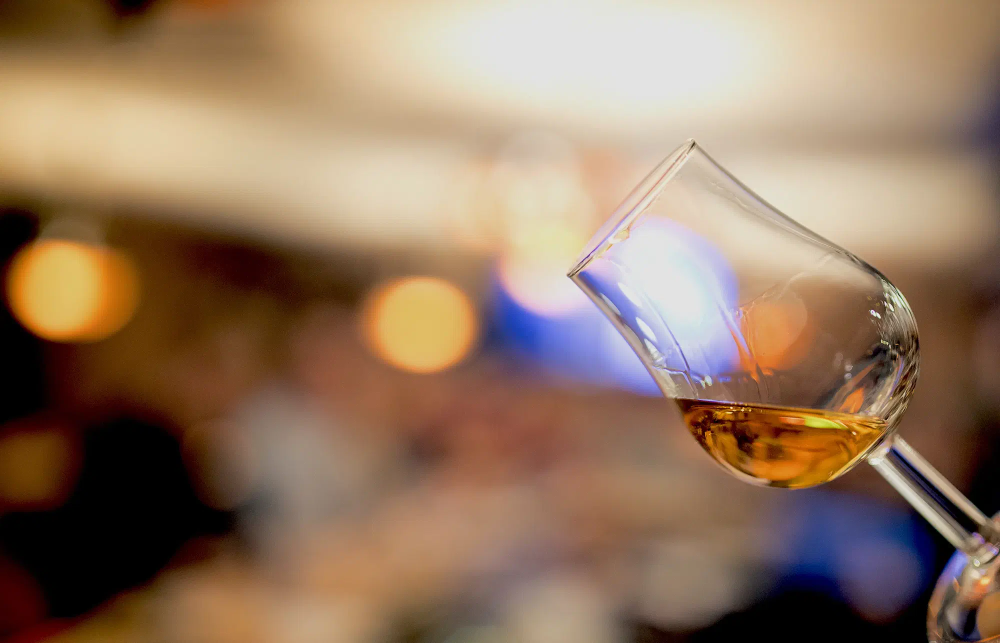Ein Nosing-Glas mit goldenem Whisky, schräg gehalten vor warmen, unscharfen Lichtern